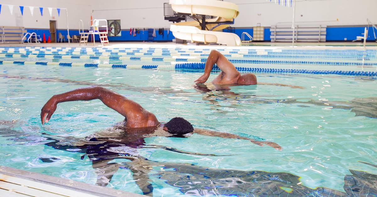 Two-people-swimming-freestyle-in-an-indoor-pool-with-lanes-and-a-water-slide-in-the-background